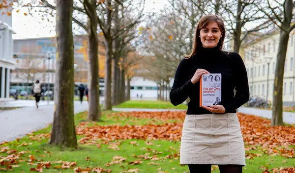 Franca Bauernfeind vor herbstlichen Bäumen auf dem Campus der Uni Erfurt. In den Händen hält sie ihr Buch.